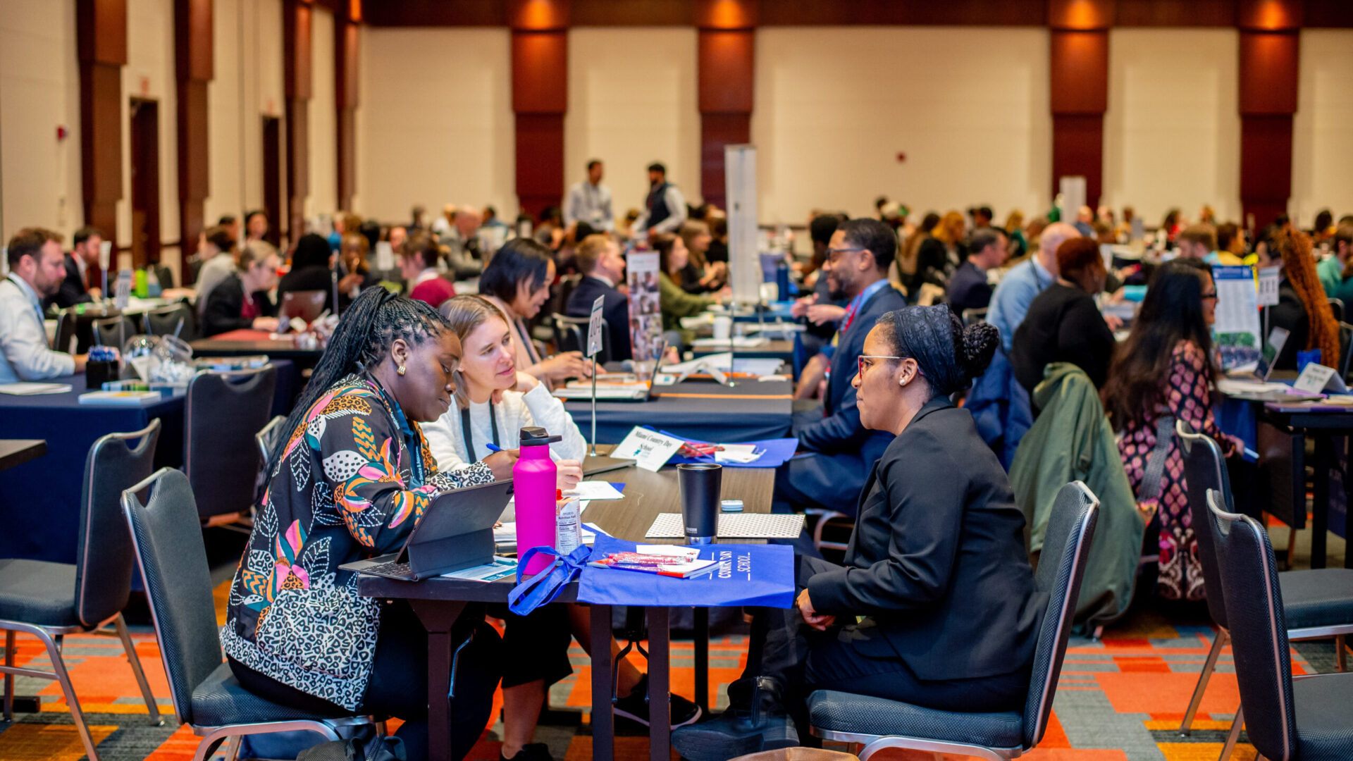 People engaged in conversations at a large networking event, seated at tables in a spacious conference room.